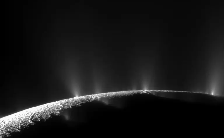Cassini’s backlit view of Enceladus shows wispy plumes rising from fractures along the moon’s curved, icy south pole into dark space.