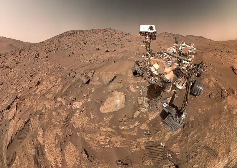 Perseverance sits beside the pale Cheyava Falls rock on Mars, with sampling marks left of the rover and wheel tracks crossing Jezero Crater’s rocky ground.
